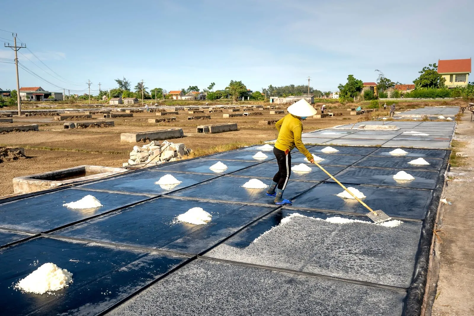 Worker in conical hat harvesting salt in the rural outdoors on a sunny day.
