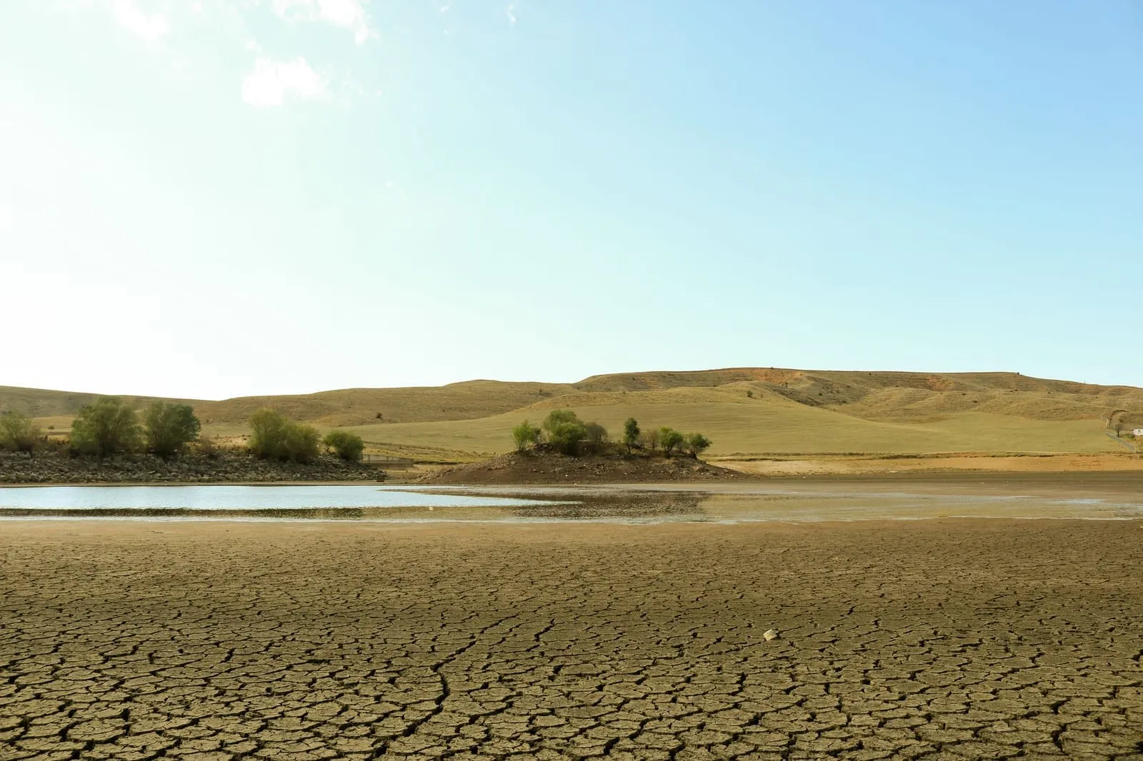 A vast arid landscape showing cracked earth, water, and sparse greenery under a clear sky.
