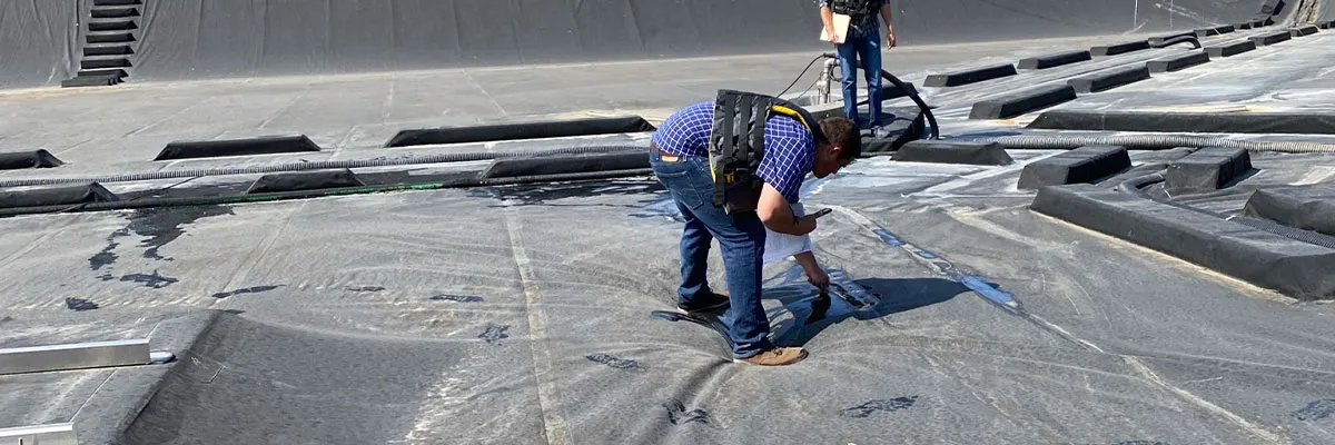 Two workers in safety harnesses kneel on a large dark geomembrane to inspect it, with pooled water on the surface