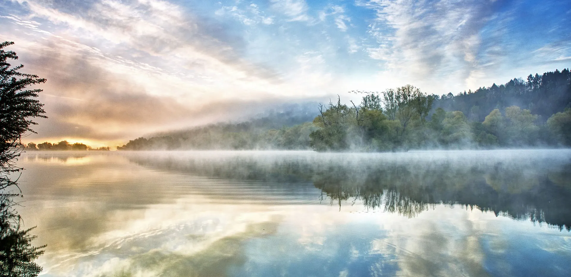 Mist drifting over a still lake at sunrise with forested hills reflected in the water