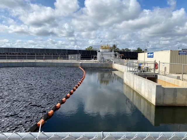 A rectangular water-treatment basin half-covered with dark floating modules, a red containment boom curving across the open water